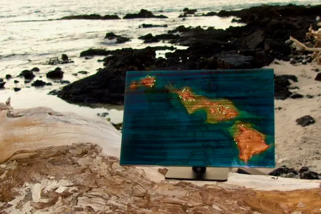 Nalu sculpture, a carved map of the Hawaiian Islands, displayed on lava rock by the shore